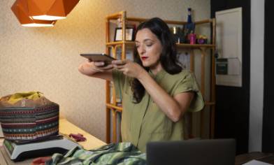 A person standing at a counter takes a photo of a patterned bag using a tablet, with folded clothes and a laptop nearby in a well-lit indoor setting