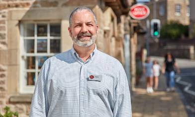 A person with a post office name badge in sharp focus in the background there is a stone building with a post office sign and three people walking on the pavement