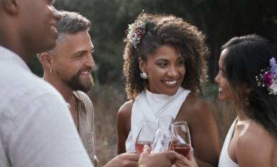 People dressed in light-coloured outfits clinking glasses of wine outdoors, with floral hair accessories visible