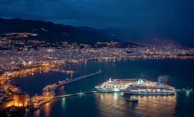 Two cruise ships are docked in a lit coastal city at night, with mountains behind 