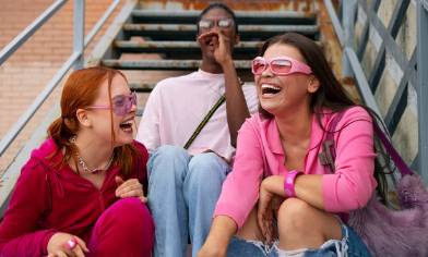 Three teens sit on outdoor stairs with brick walls and railings behind them. They wear red, white and pink outfits and sunglasses
