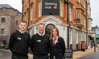 Three people in black uniforms stand outside a red brick building labelled 'Banking Hub' with white trim and large windows. It proudly stands on a high street