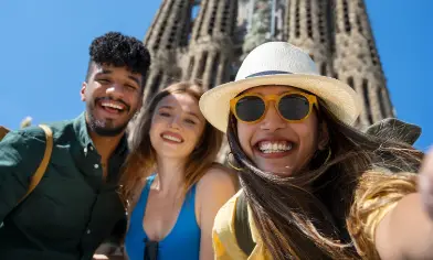 Three people take a selfie in front of the Sagrada Familia in Barcelona, with the basilica’s ornate towers rising behind them under a clear blue sky