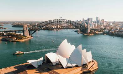 Sydney opera house in the foreground with Sydney Harbour Bridge beyond and a city skyline further back still. A ferry crosses the water between the opera house and the bridge