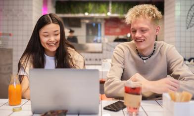 Two smiling people are hanging out at a café table with snacks and drinks, looking at a laptop. The surrounding walls and tabletop are made of white tiles
