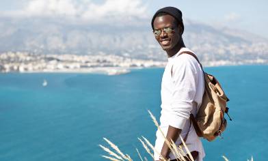 A person wearing a backpack, cap and sunglasses, standing on a cliffside overlooking a bay