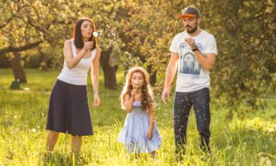 A family stands in a sunlit meadow blowing dandelion seeds surrounded by tall grass and trees