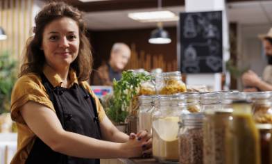 A person in a black apron stands behind a counter displaying jars of food, with a chalkboard and other people in shop in the background