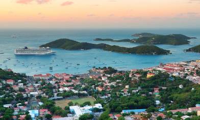 Aerial view of a coastal town with a large cruise ship docked near small islands, surrounded by buildings, roads, green spaces and boats, under an orange and pink sky 