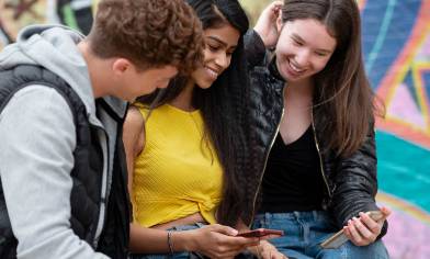Three teens sit in front of a bright graffiti wall, checking something out on a phone together 
