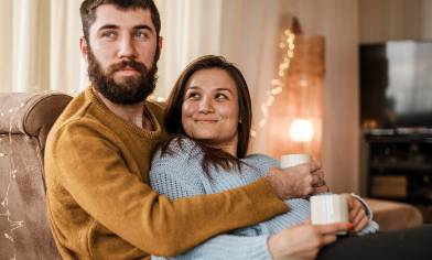 A couple sit at home, arms around each other and holding coffee cups