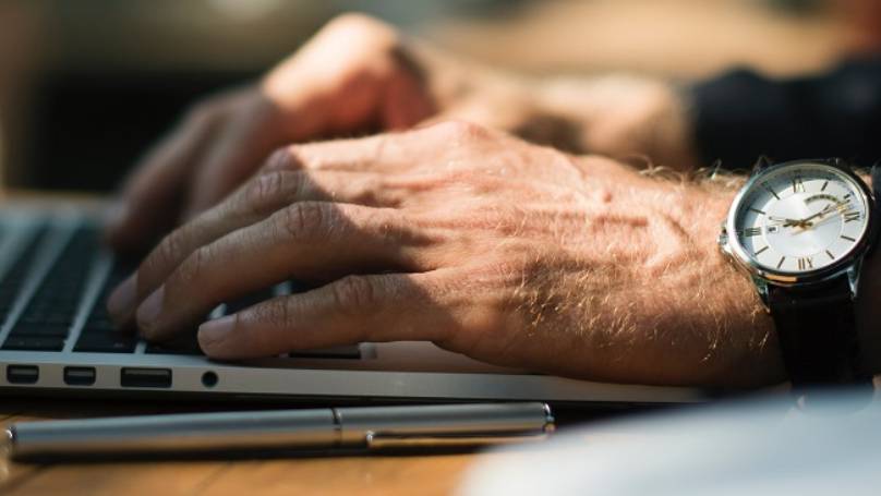 A man's hand, wearing a watch, typing on his laptop on his desk