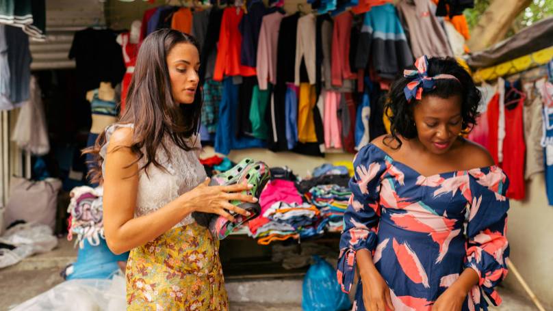 Two individuals standing in a clothing market with colourful garments.