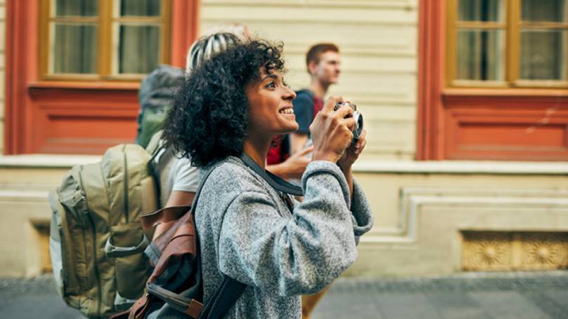Woman wearing backpack holding a camera taking a photo in a street