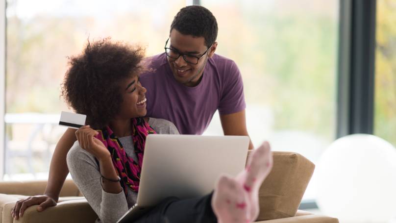 Woman and partner laughing together, whilst she is sitting down with laptop, holding a credit card in one hand