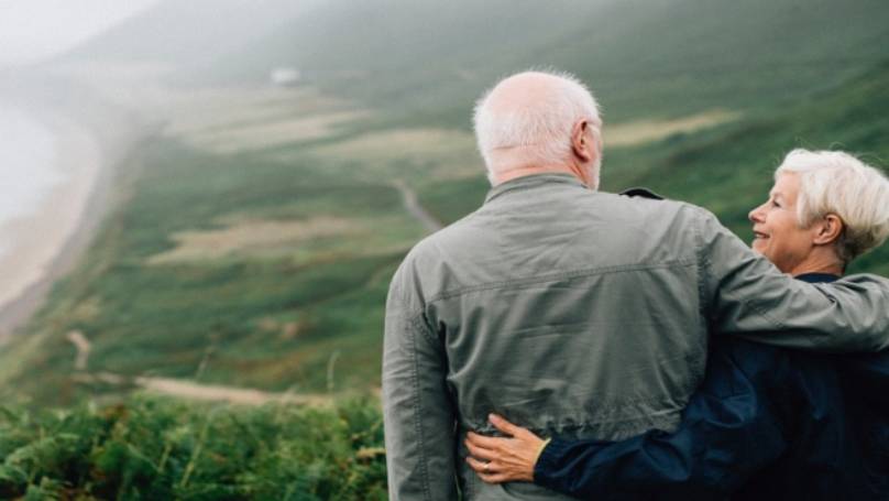 Elderly couple looking at each other lovingly, stood on a high hillside overlooking coastline below