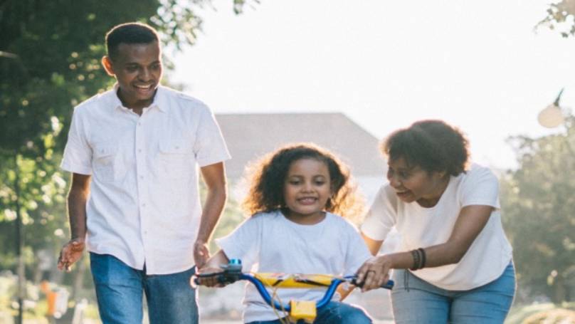 Mother and father teaching their little girl to ride a bike along a sunlit road