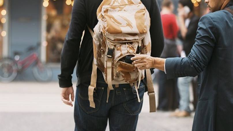 Man with rolled sleeve taking item from backpack on another man walking in ahead in a street