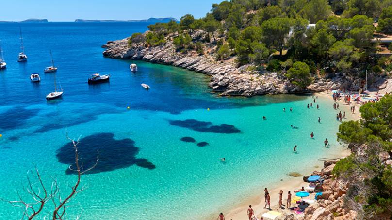 Rocky and green coastline with people on sandy beach surrounded with blue waters and boats