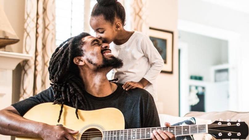 Little girl kissing her daddy on the forehead while he's playing a guitar, in their home