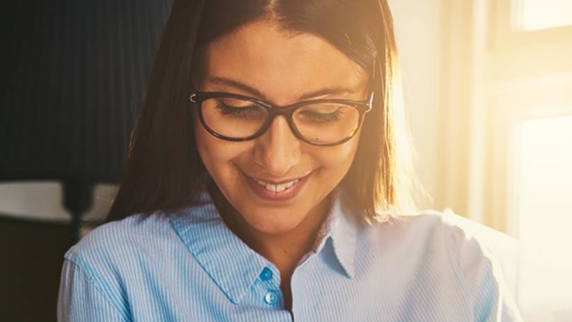 Young smiling woman in light blue shirt wearing glasses, sat in a sunlit lounge, looking at laptop