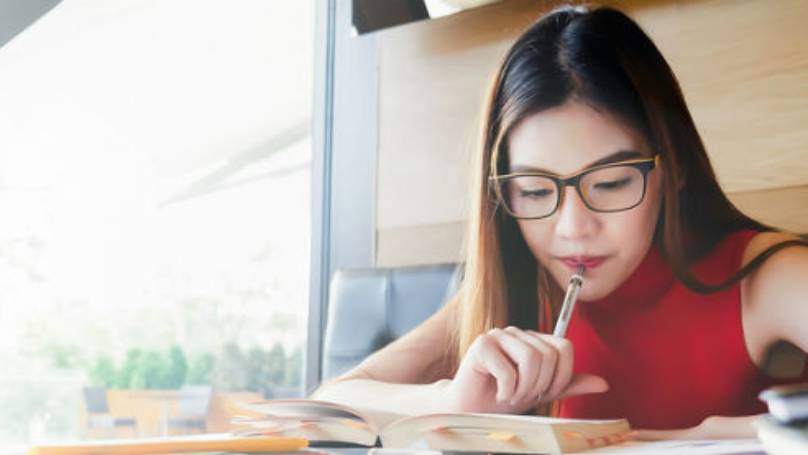 Woman sat in a well-lit café, holding pen to her mouth while reading from a book for her studies
