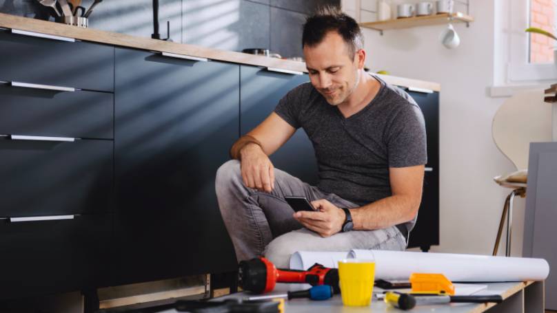 Man kneeling down on one knee looking at his phone with DIY tools in front of him
