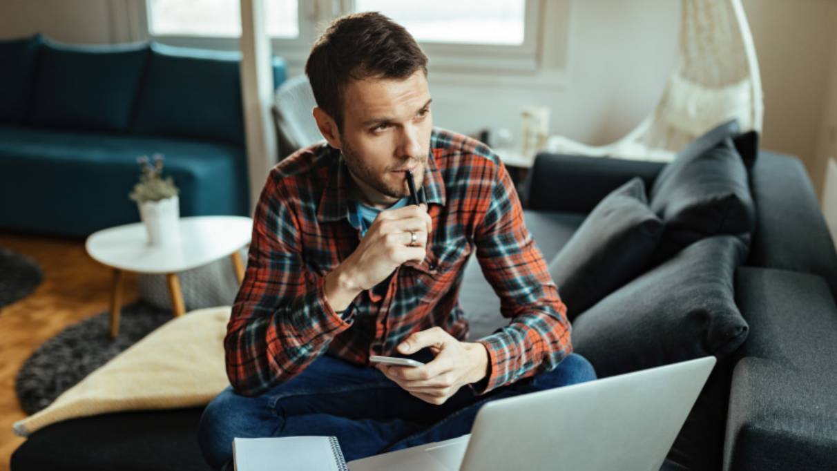 Man sitting in front of laptop, looking thoughtful, holding a pen and phone either hand