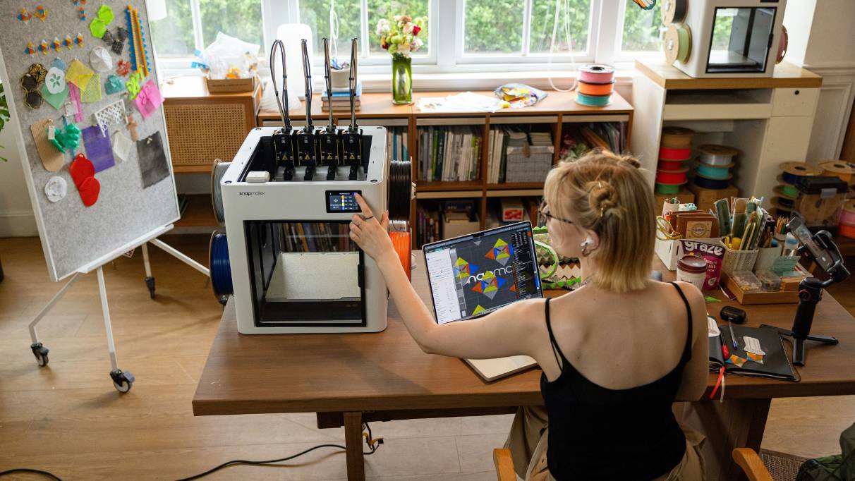 A person seated at a desk in a craft room, operating a 3D printer.