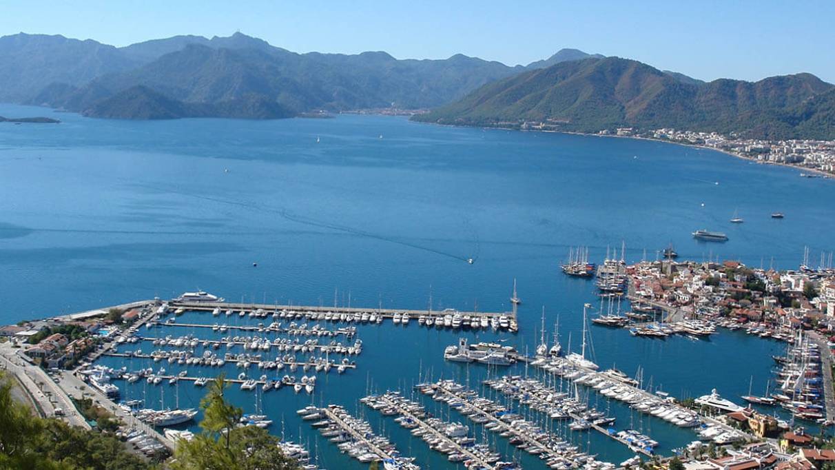 a marina with rows of boats and yachts docked along a calm coastline, surrounded by mountainous terrain and nearby buildings