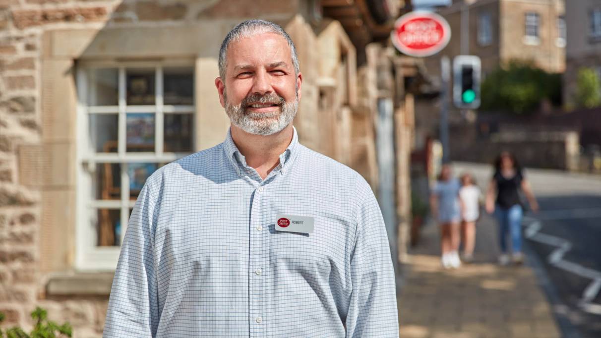 A person with a post office name badge in sharp focus in the background there is a stone building with a post office sign and three people walking on the pavement