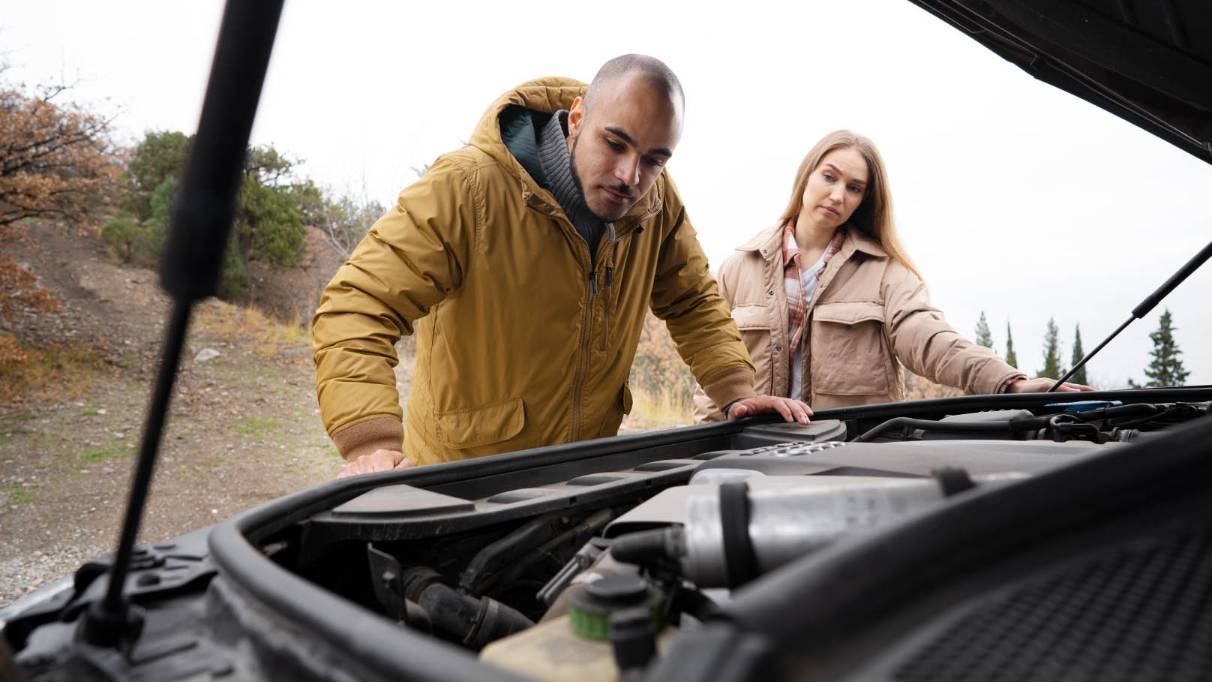 A couple stands by the open bonnet of a presumably broken-down car, looking down at the engine inside