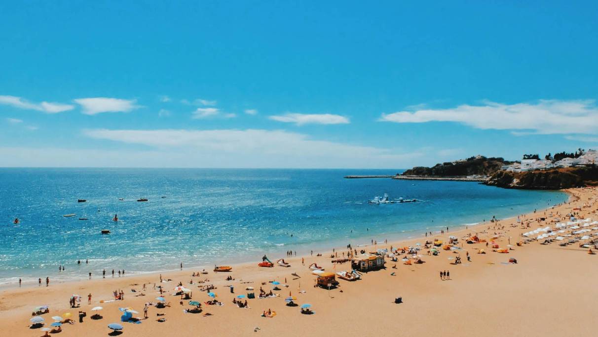 Raised view of a sandy beach where a light sea and sky. The beach is dotted with umbrellas, blankets and people enjoying their holidays. In the distance, small white buildings and dark vegetation line the shore. 