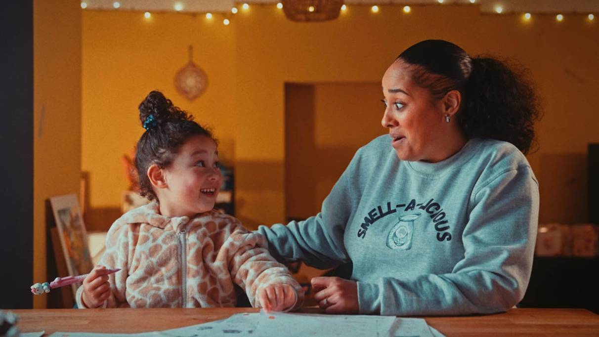 A child holding a pencil sits at a table with an adult wearing a 'SMELL-A-LICIOUS' sweatshirt