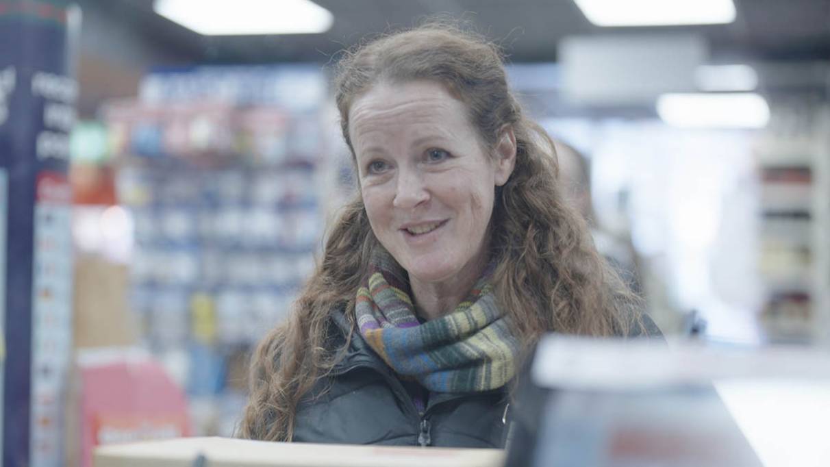 A person standing at the counter in a brightly lit Post Office branch, with shelves of products in the background