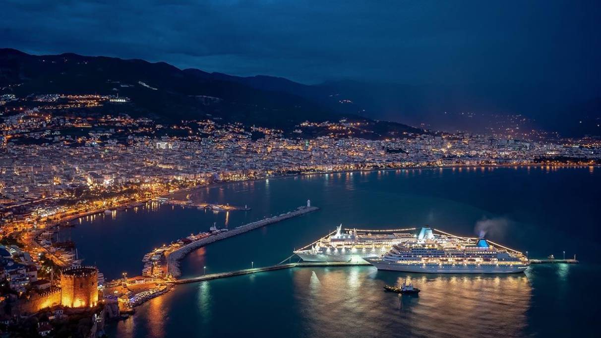 Two cruise ships are docked in a lit coastal city at night, with mountains behind 