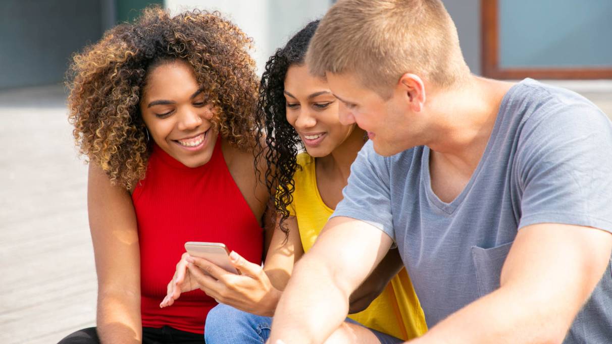 Three young friends in bright clothes smile at something on one of their phones while sitting on the steps of their university building.