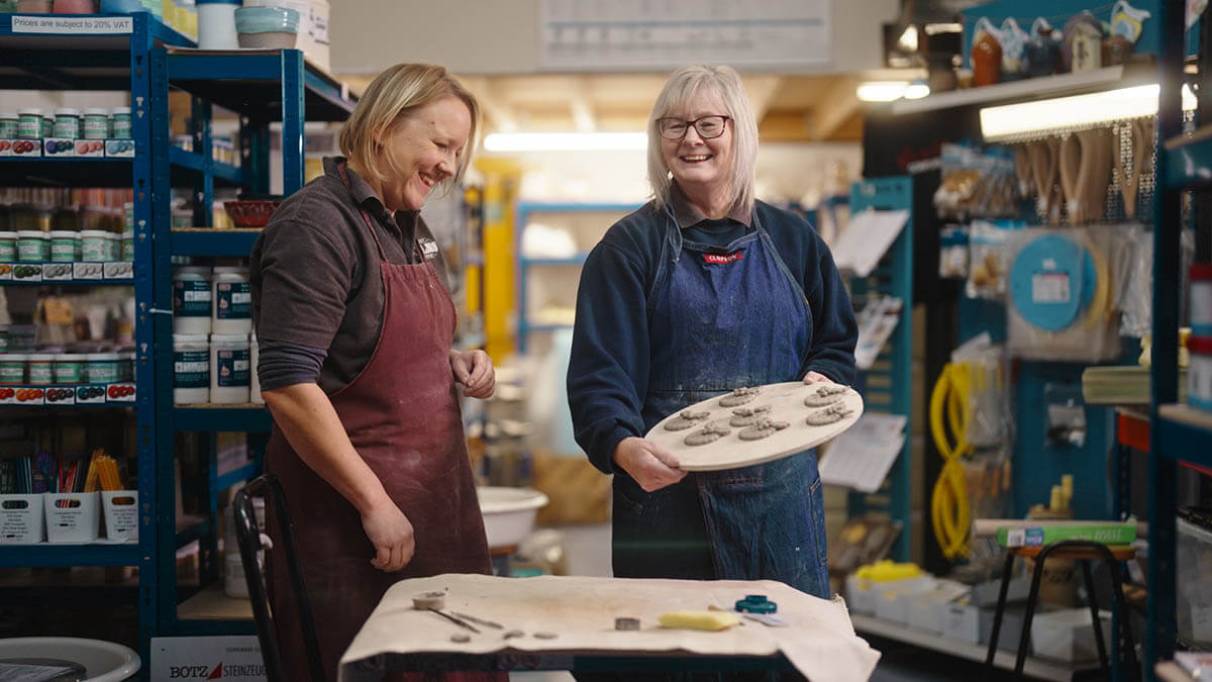 Two people in a workshop wearing aprons, one holding a tray of small clay objects, surrounded by shelves filled with tools and supplies 