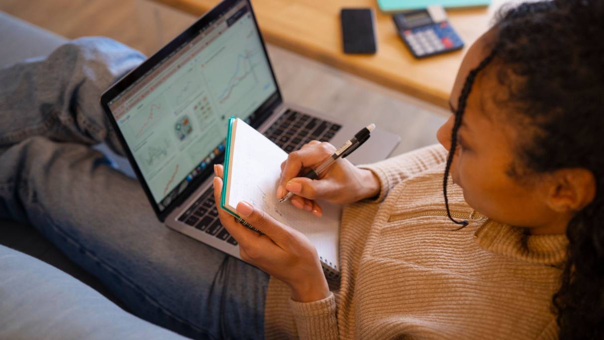 Person sitting on a sofa writing in a notebook while using a laptop, with charts visible on the screen; a smartphone, calculator, and folder are on the table nearby.
