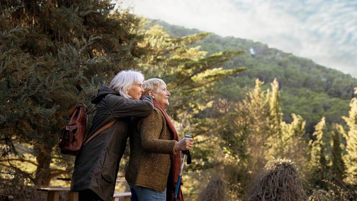 Two people stand together in a forested area, with one person hugging the other from behind while the other holds a walking stick