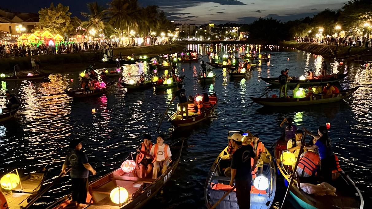 Boats with lanterns float on a river at night, with crowds watching from lit-up riverbanks.