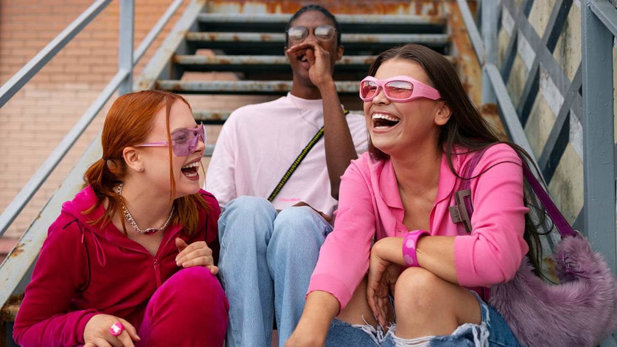 Three teens sit on outdoor stairs with brick walls and railings behind them. They wear red, white and pink outfits and sunglasses