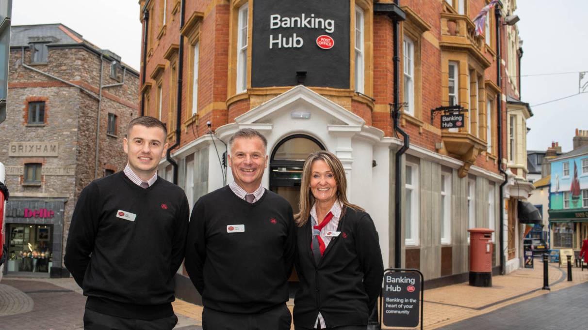Three people in black uniforms stand outside a red brick building labelled 'Banking Hub' with white trim and large windows. It proudly stands on a high street