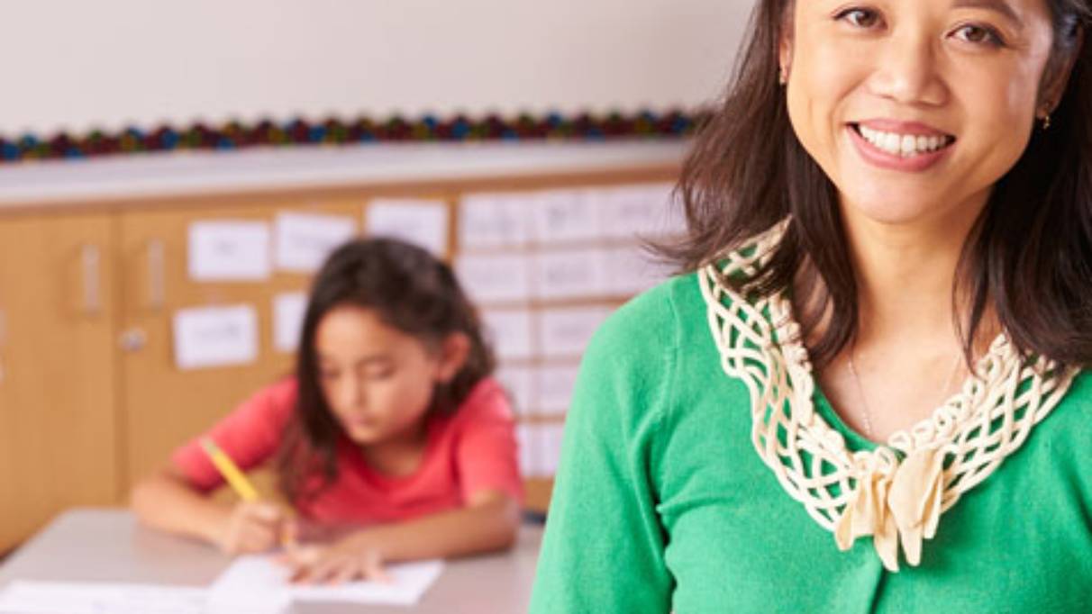 Smiling female teacher in green cardigan in foreground, child student writing in background