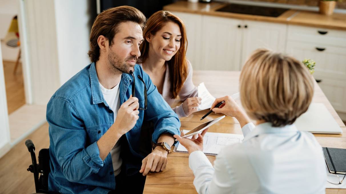 Three people at a kitchen table, one in a wheelchair holding a pen, discussing documents. One holds out a digital tablet for the others to read