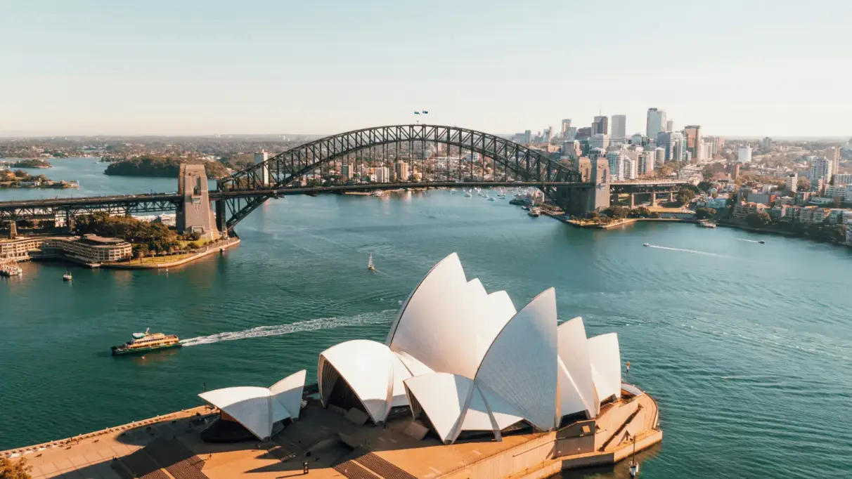 Sydney opera house in the foreground with Sydney Harbour Bridge beyond and a city skyline further back still. A ferry crosses the water between the opera house and the bridge