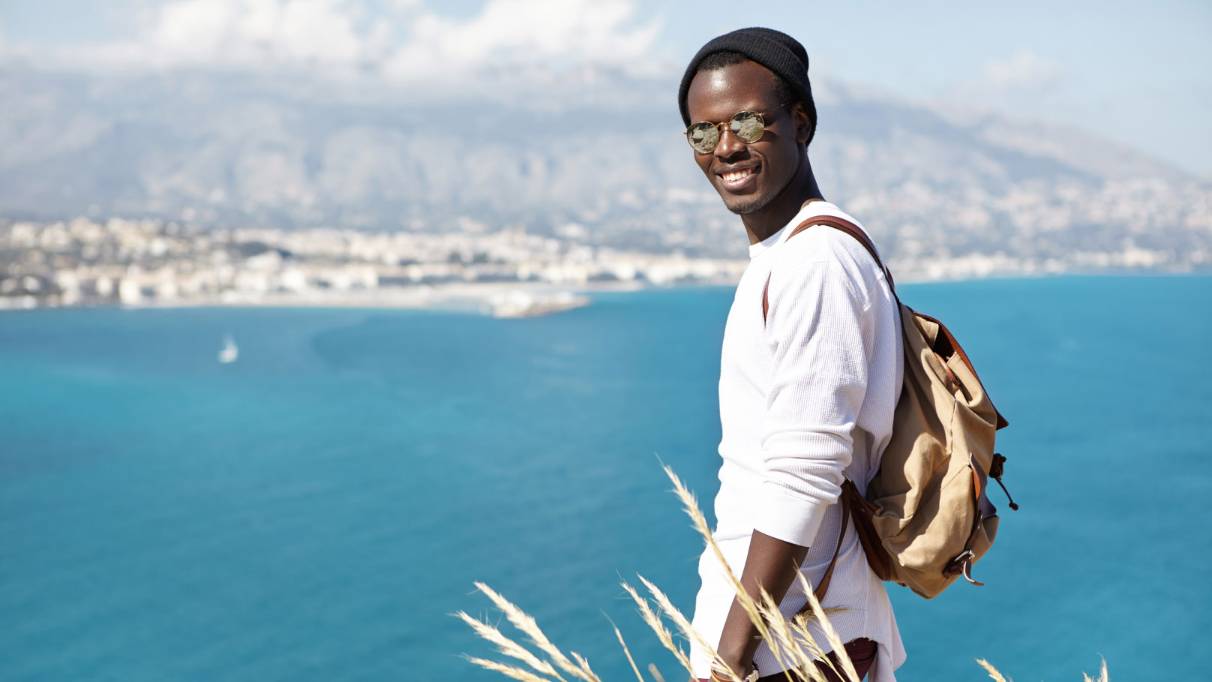 A person wearing a backpack, cap and sunglasses, standing on a cliffside overlooking a bay