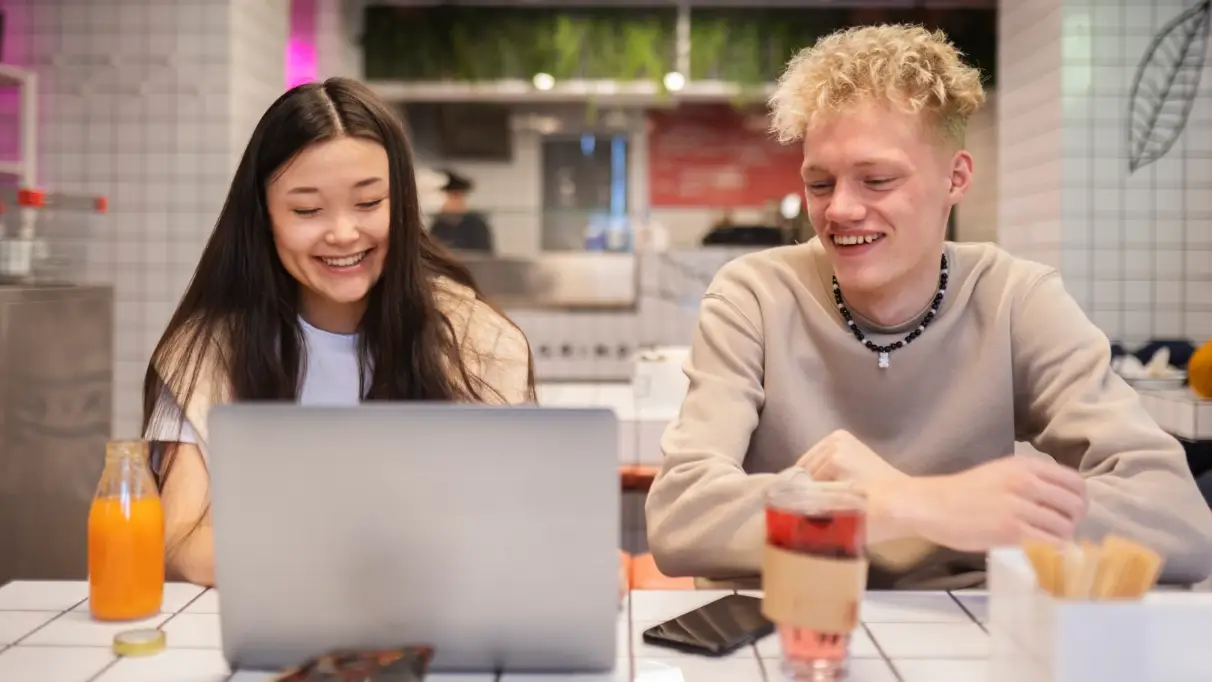Two smiling people are hanging out at a café table with snacks and drinks, looking at a laptop. The surrounding walls and tabletop are made of white tiles