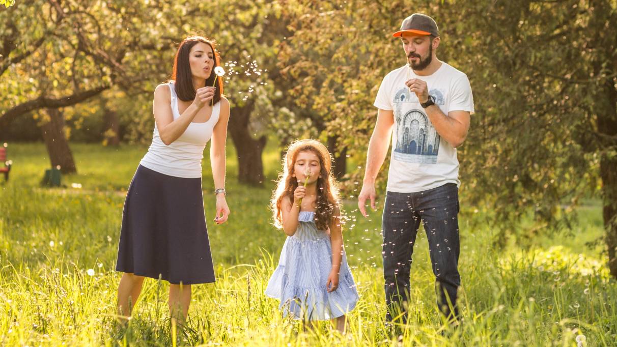 A family stands in a sunlit meadow blowing dandelion seeds surrounded by tall grass and trees