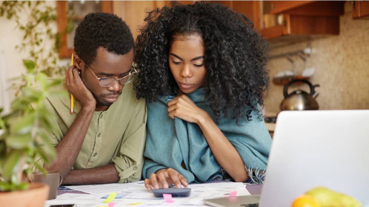 Two people looking at bills on a kitchen table with a laptop, calculator and various documents